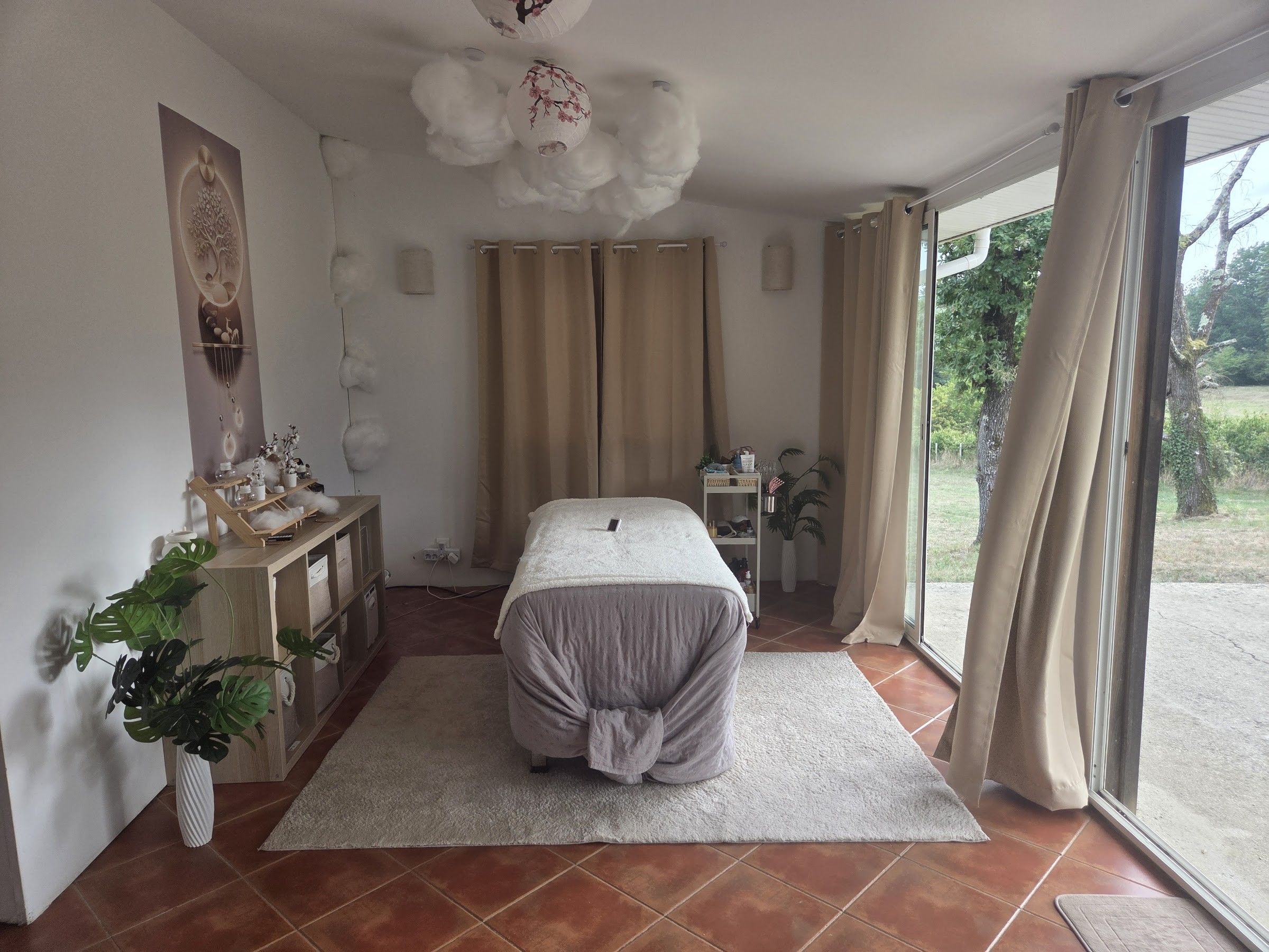 La salle de soin de Lumaïa en journée : table de soin, plafond de nuages et baie vitrée ouverte sur le jardin